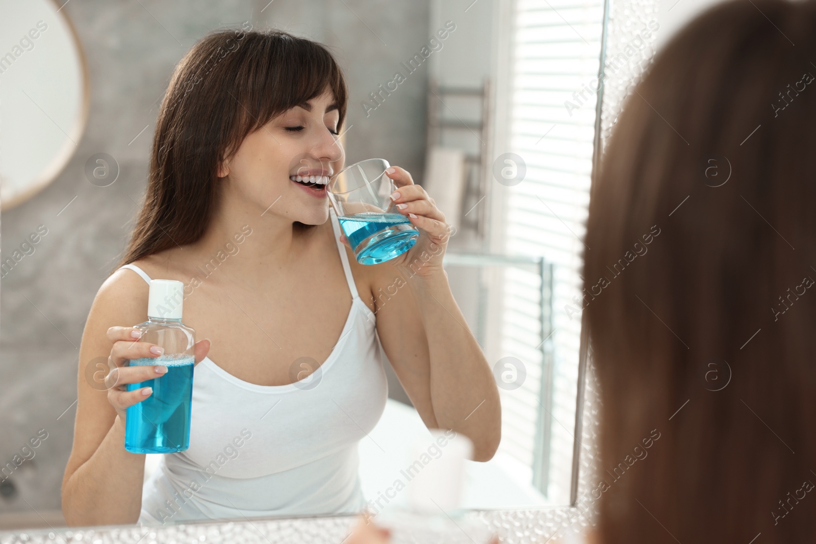 Photo of Young woman using mouthwash near mirror in bathroom