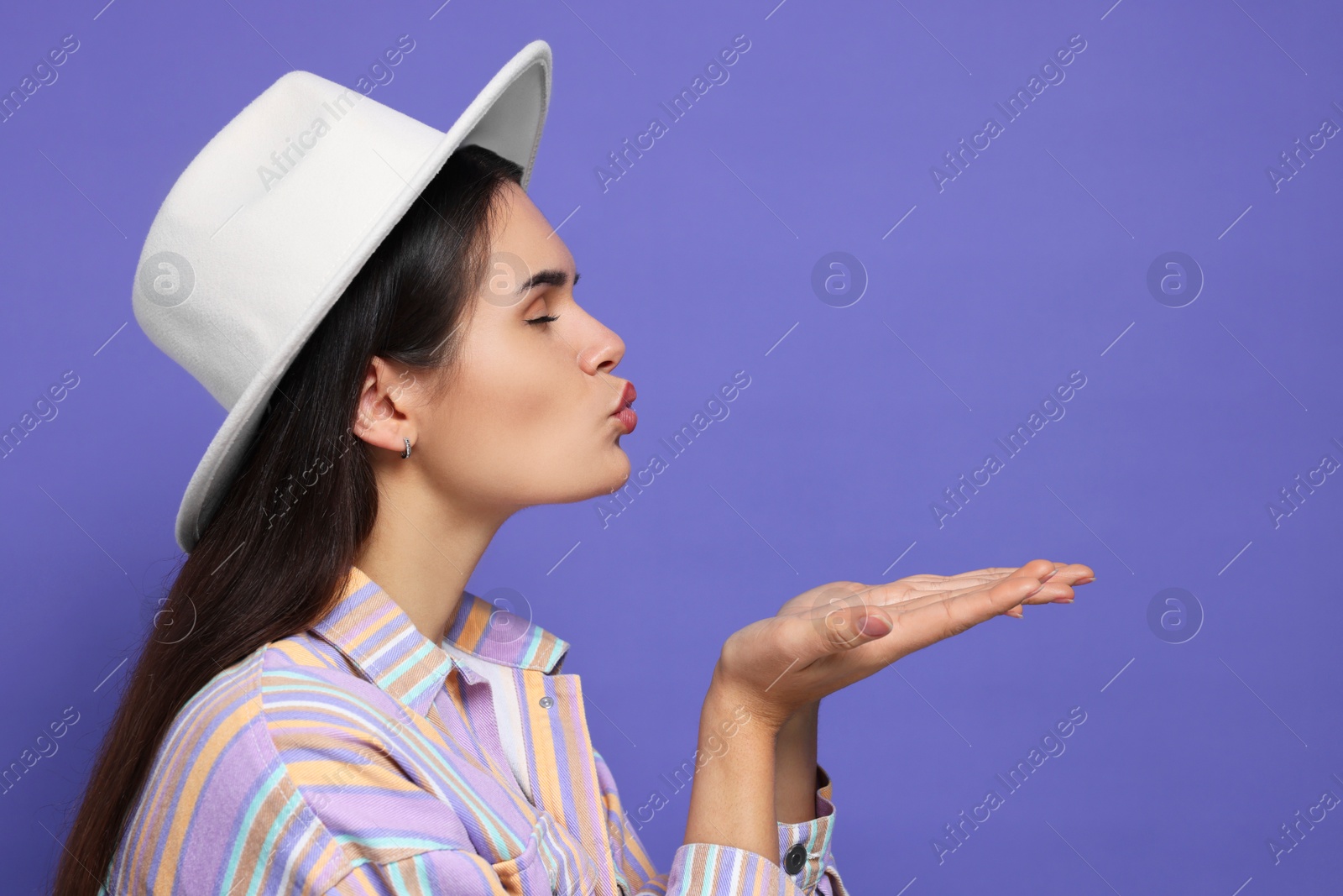 Photo of Beautiful young woman with stylish hat blowing kiss on purple background. Space for text