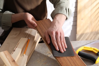 Man polishing wooden plank with sandpaper at grey table indoors, closeup