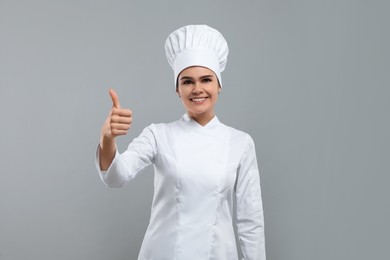 Happy female chef wearing uniform and cap showing thumbs up on light grey background