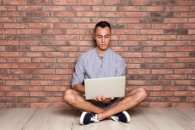 Young man sitting on floor with laptop against brick wall