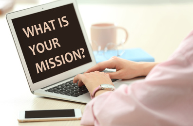 Image of Young woman using modern laptop with question WHAT IS YOUR MISSION? on screen indoors, closeup