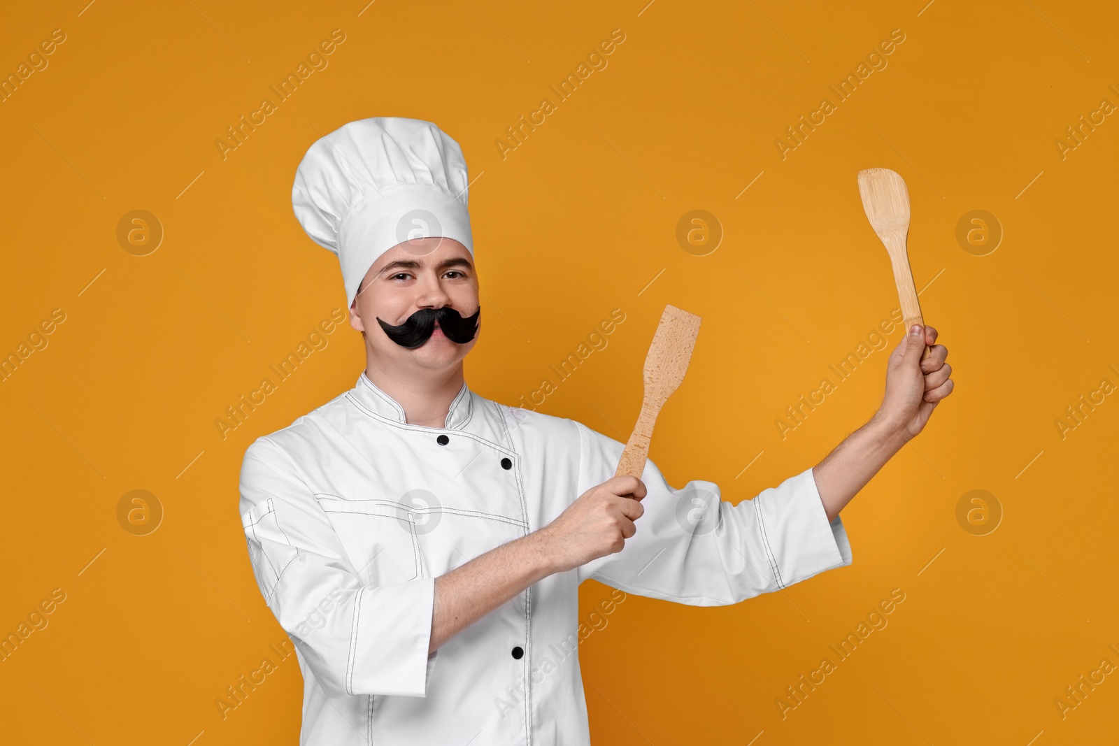 Photo of Portrait of happy confectioner with funny artificial moustache holding wooden spatulas on orange background