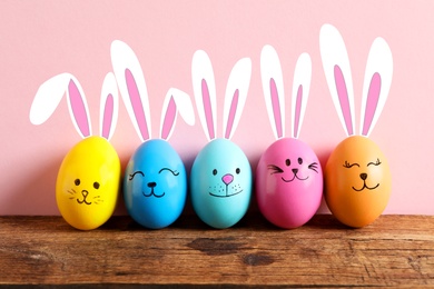 Colorful eggs as Easter bunnies on wooden table against pink background