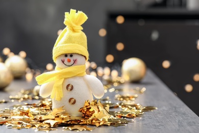 Photo of Snowman toy and confetti stars on grey table against blurred festive lights, space for text. Christmas decoration