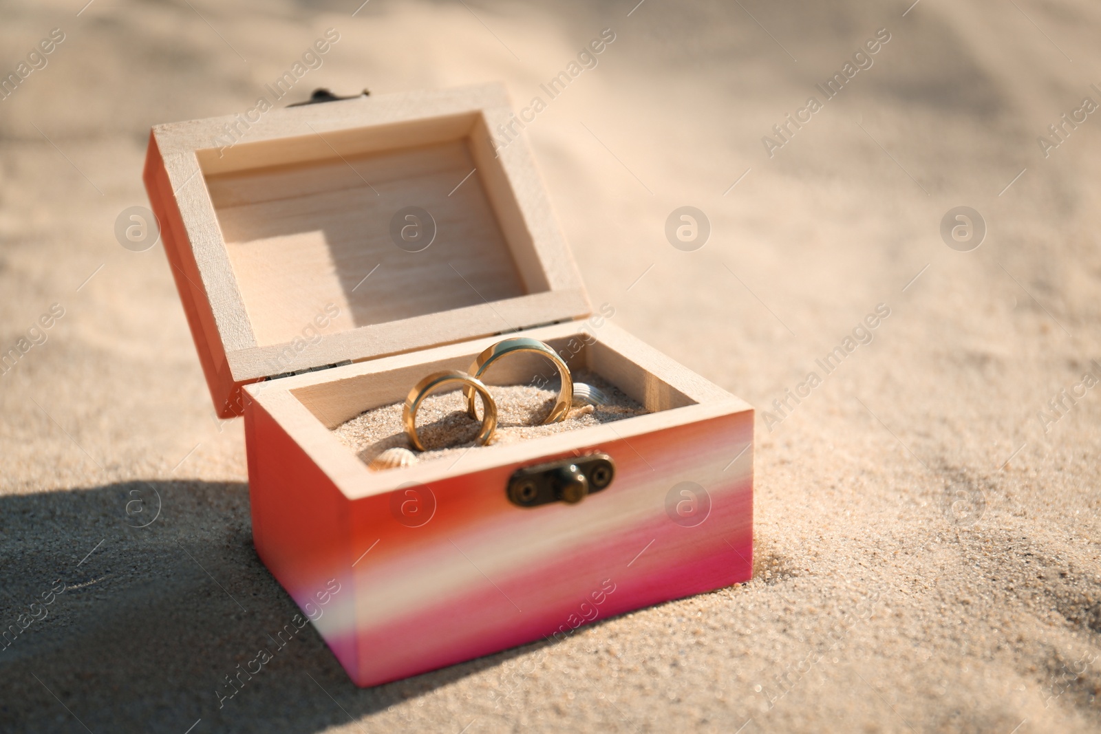 Image of Wooden box in color of lesbian flag with golden wedding rings on sandy beach