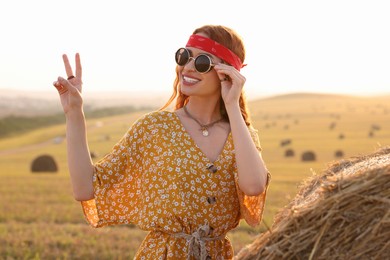 Happy hippie woman showing peace sign near hay bale in field