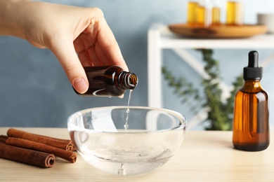 Photo of Woman adding cinnamon essential oil from bottle into bowl of water on table indoors, closeup