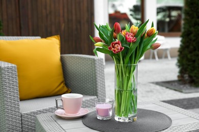 Photo of Beautiful bouquet of colorful tulips, candle and cup with drink on rattan garden table outdoors