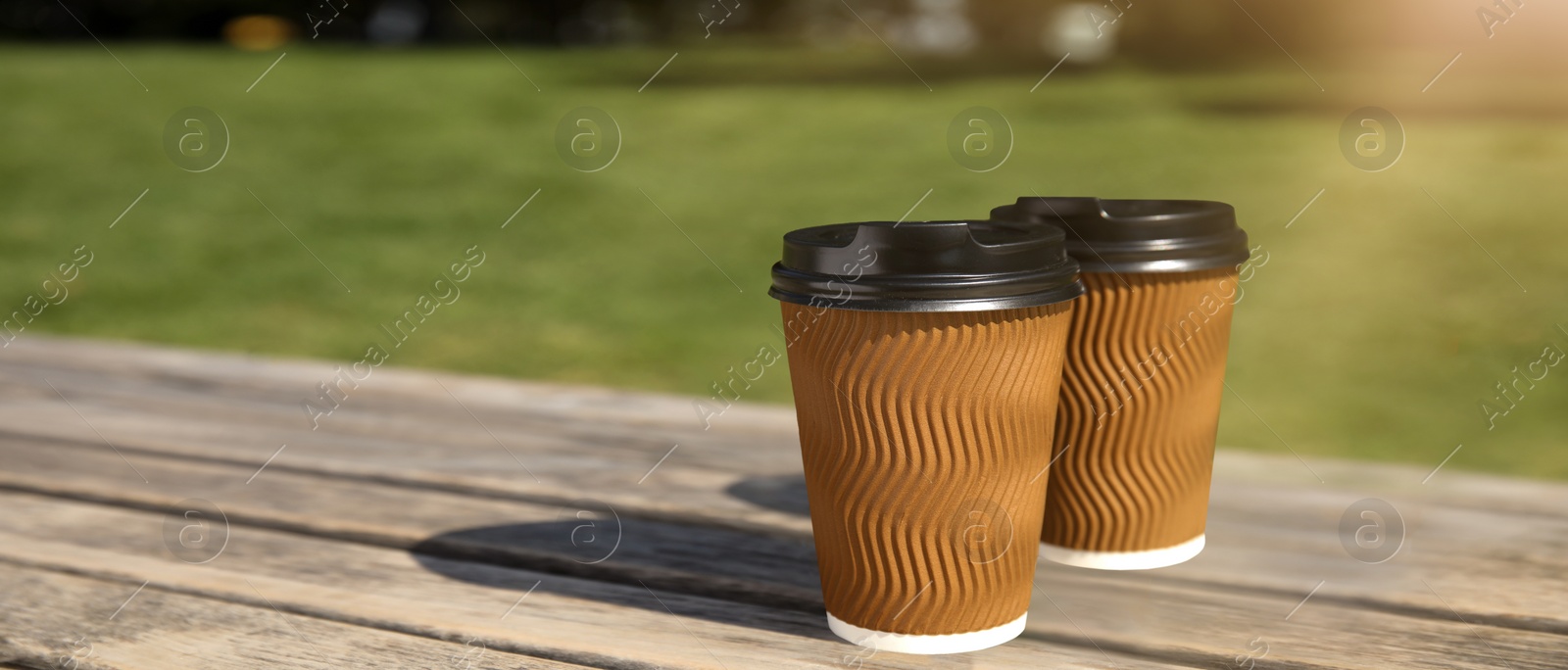 Image of Takeaway cardboard coffee cups with plastic lids on wooden bench outdoors, space for text. Banner design