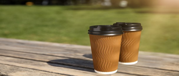 Image of Takeaway cardboard coffee cups with plastic lids on wooden bench outdoors, space for text. Banner design