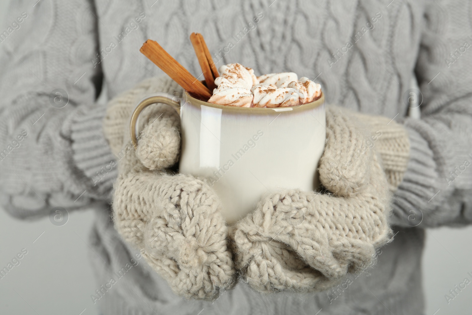 Photo of Woman in knitted mittens holding cup of delicious hot chocolate with marshmallows and cinnamon sticks, closeup