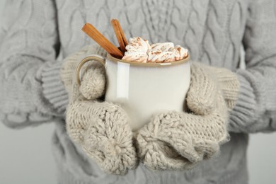 Photo of Woman in knitted mittens holding cup of delicious hot chocolate with marshmallows and cinnamon sticks, closeup