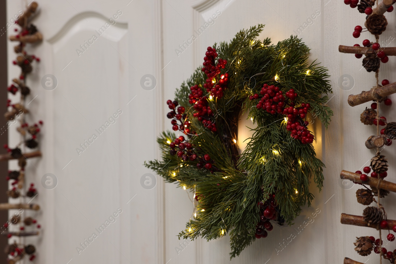 Photo of Beautiful Christmas wreath with red berries and fairy lights hanging on white door, space for text