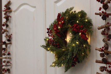 Photo of Beautiful Christmas wreath with red berries and fairy lights hanging on white door, space for text