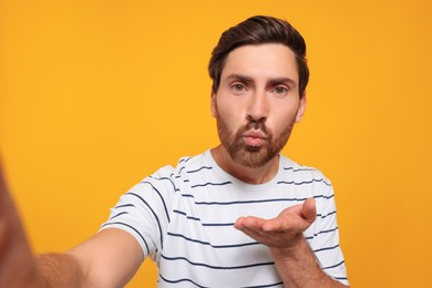 Handsome man blowing kiss while taking selfie on orange background. Space for text