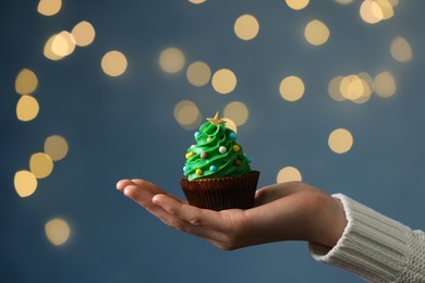 Photo of Woman holding tasty Christmas cupcake against blurred festive lights, closeup