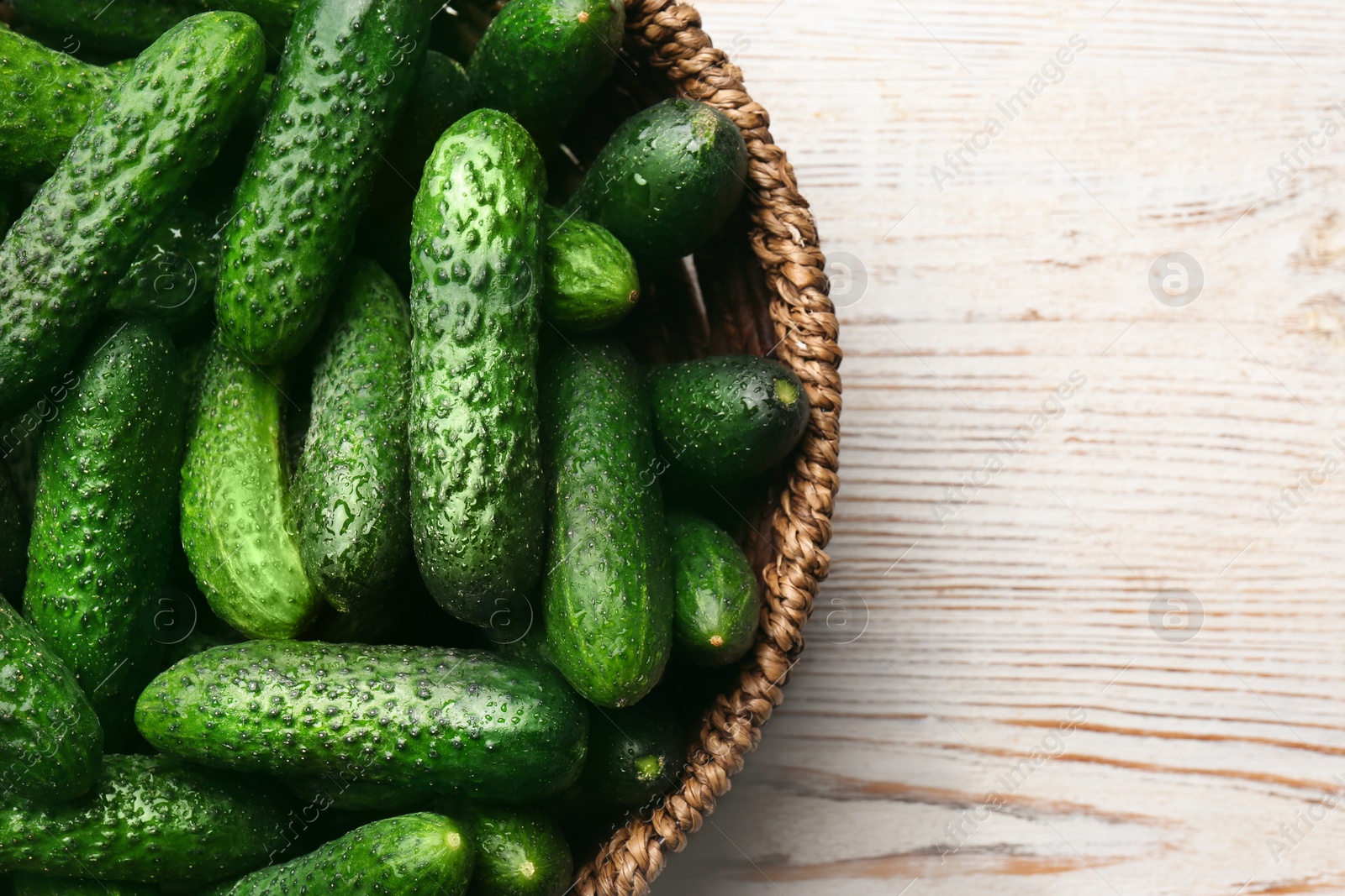 Photo of Fresh ripe cucumbers in wicker basket on white wooden table, top view. Space for text