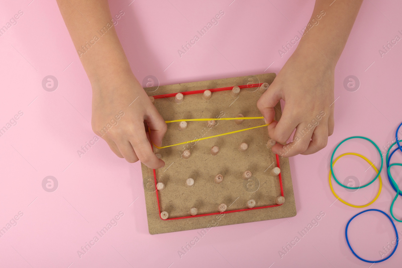 Photo of Motor skills development. Girl playing with geoboard and rubber bands at pink table, top view