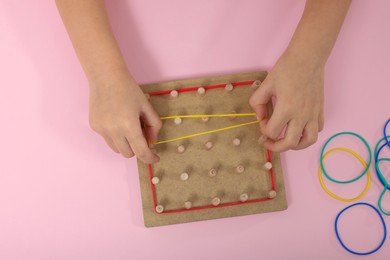Photo of Motor skills development. Girl playing with geoboard and rubber bands at pink table, top view