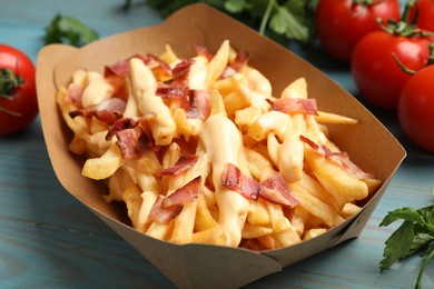 Photo of Tasty potato fries, cheese sauce, bacon and products in paper container on light blue wooden table, closeup