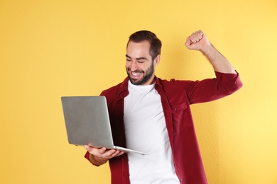 Emotional young man with laptop celebrating victory on color background