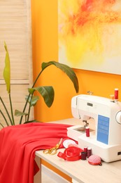 Photo of Modern sewing machine with cloth and craft accessories on wooden table near orange wall in room