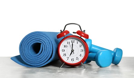 Alarm clock, yoga mat and dumbbells on marble table against grey background. Morning exercise