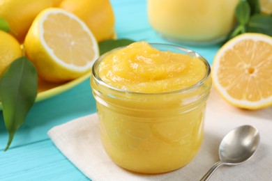 Photo of Delicious lemon curd in glass jar, fresh citrus fruits and spoon on light blue wooden table, closeup