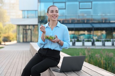 Smiling businesswoman with lunch box sitting near laptop outdoors