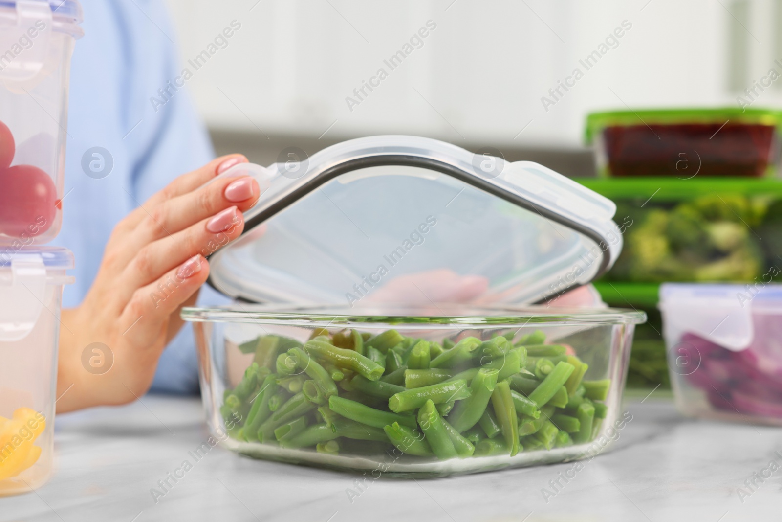 Photo of Woman sealing container with green beans at white marble table in kitchen, closeup. Food storage