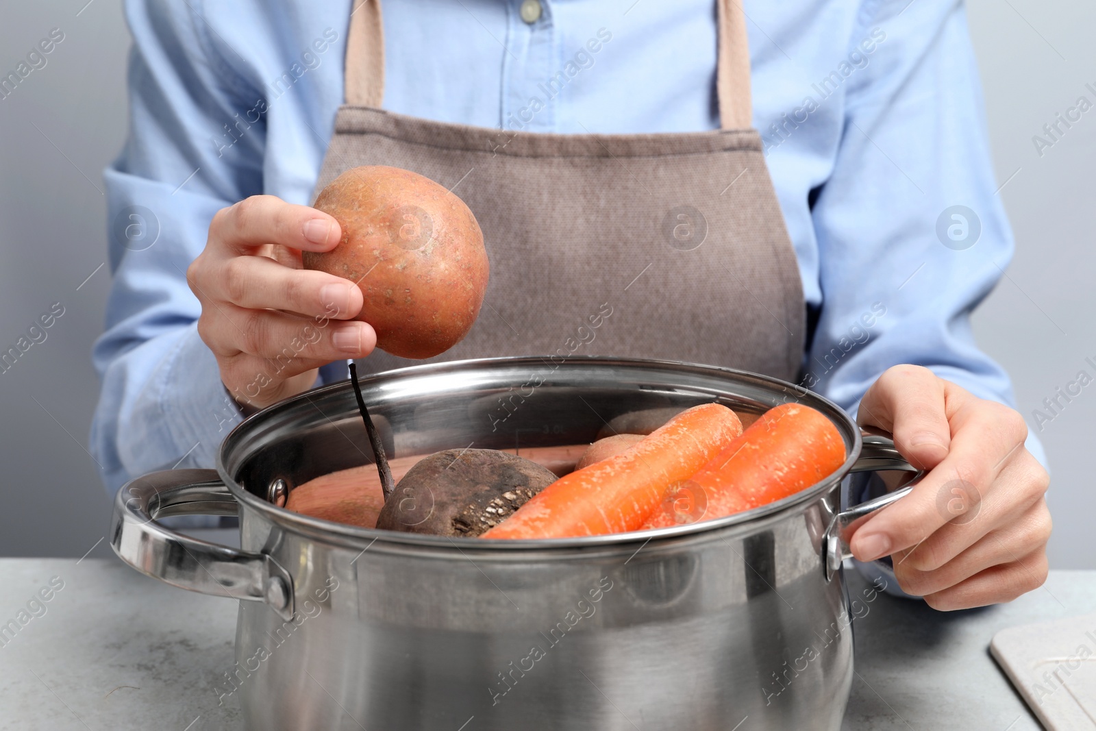 Photo of Woman putting potato into pot with fresh vegetables at white table, closeup. Cooking vinaigrette salad