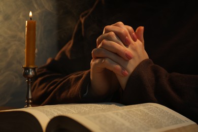 Photo of Woman praying at table with burning candle and Bible, closeup