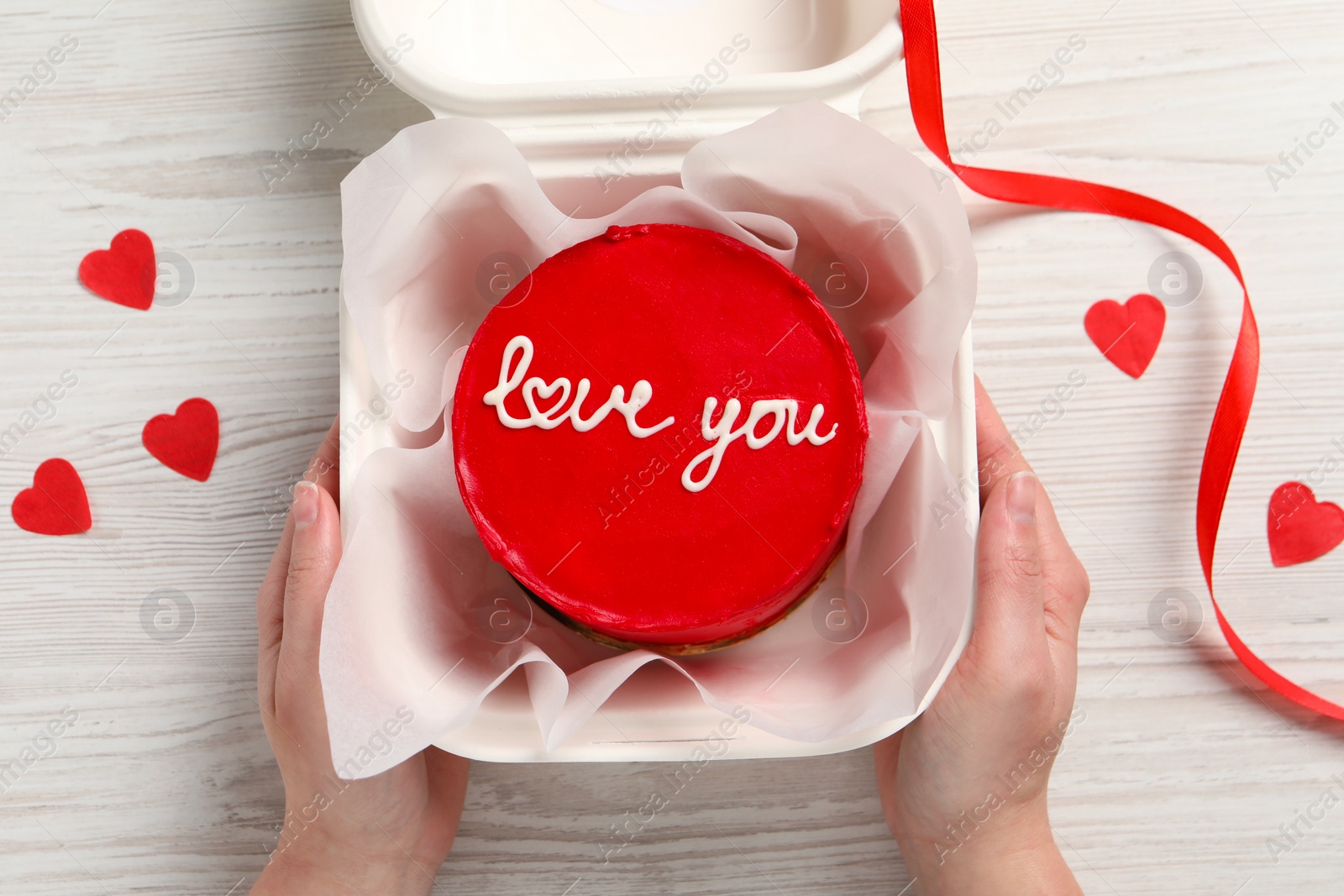 Photo of Woman holding takeaway box with bento cake at white wooden table, top view. St. Valentine's day surprise