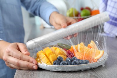 Photo of Woman putting plastic food wrap over plate of fresh fruits and berries at wooden table indoors, closeup