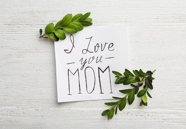 Photo of Paper sheet with words I LOVE YOU MOM and green branches on white wooden table, flat lay