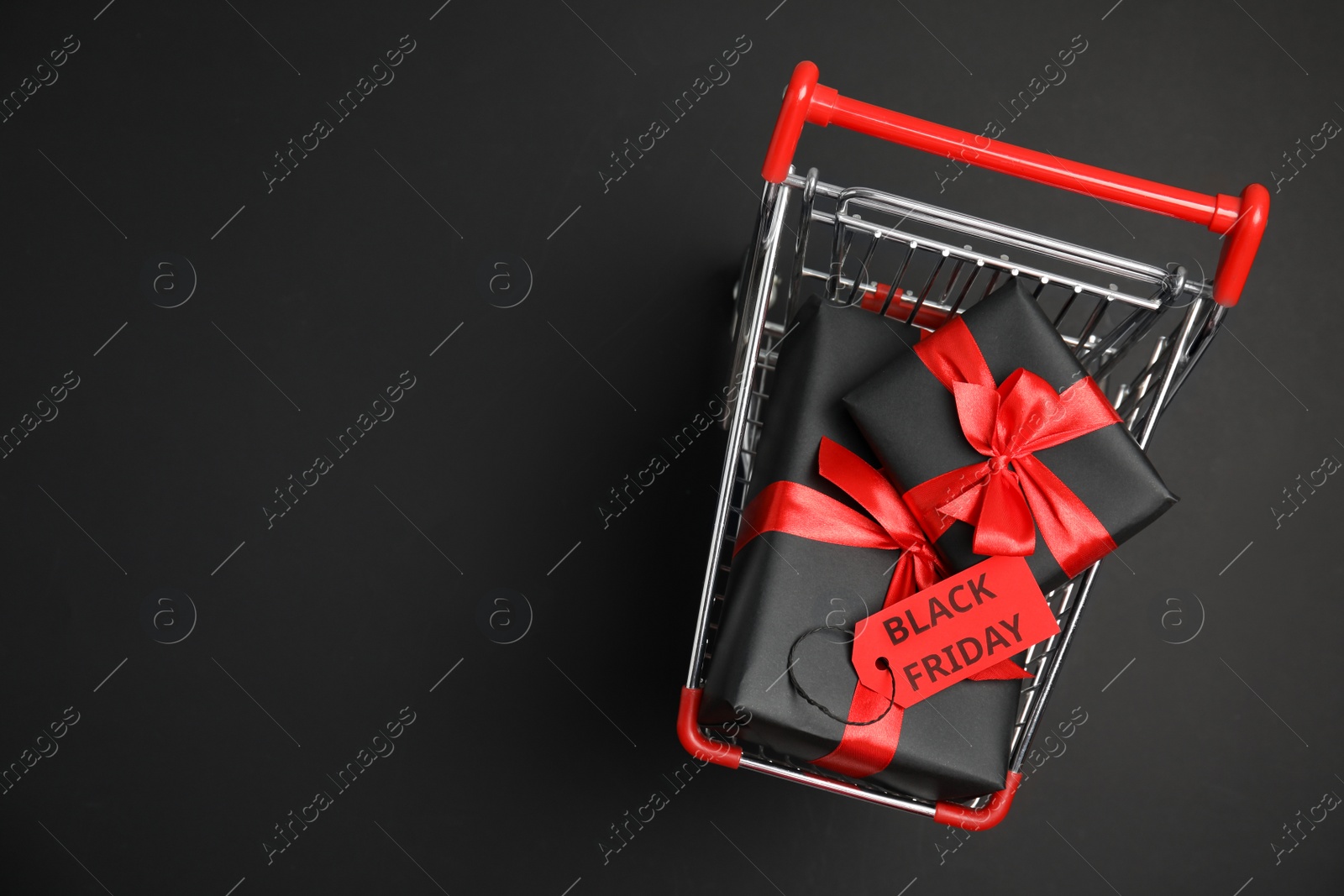 Photo of Small shopping cart with wrapped gift boxes and space for text on dark background, top view. Black Friday sale