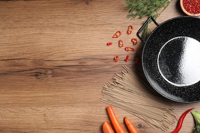 Empty iron wok surrounded by raw ingredients on wooden table, flat lay. Space for text