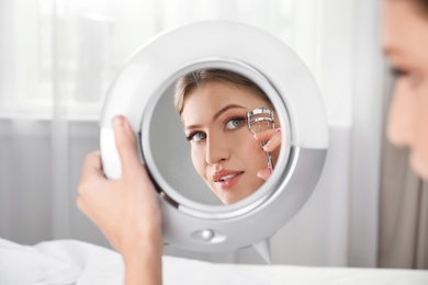 Photo of Young woman with long eyelashes and curler looking in mirror indoors