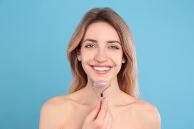 Young woman using natural rose quartz face roller on light blue background