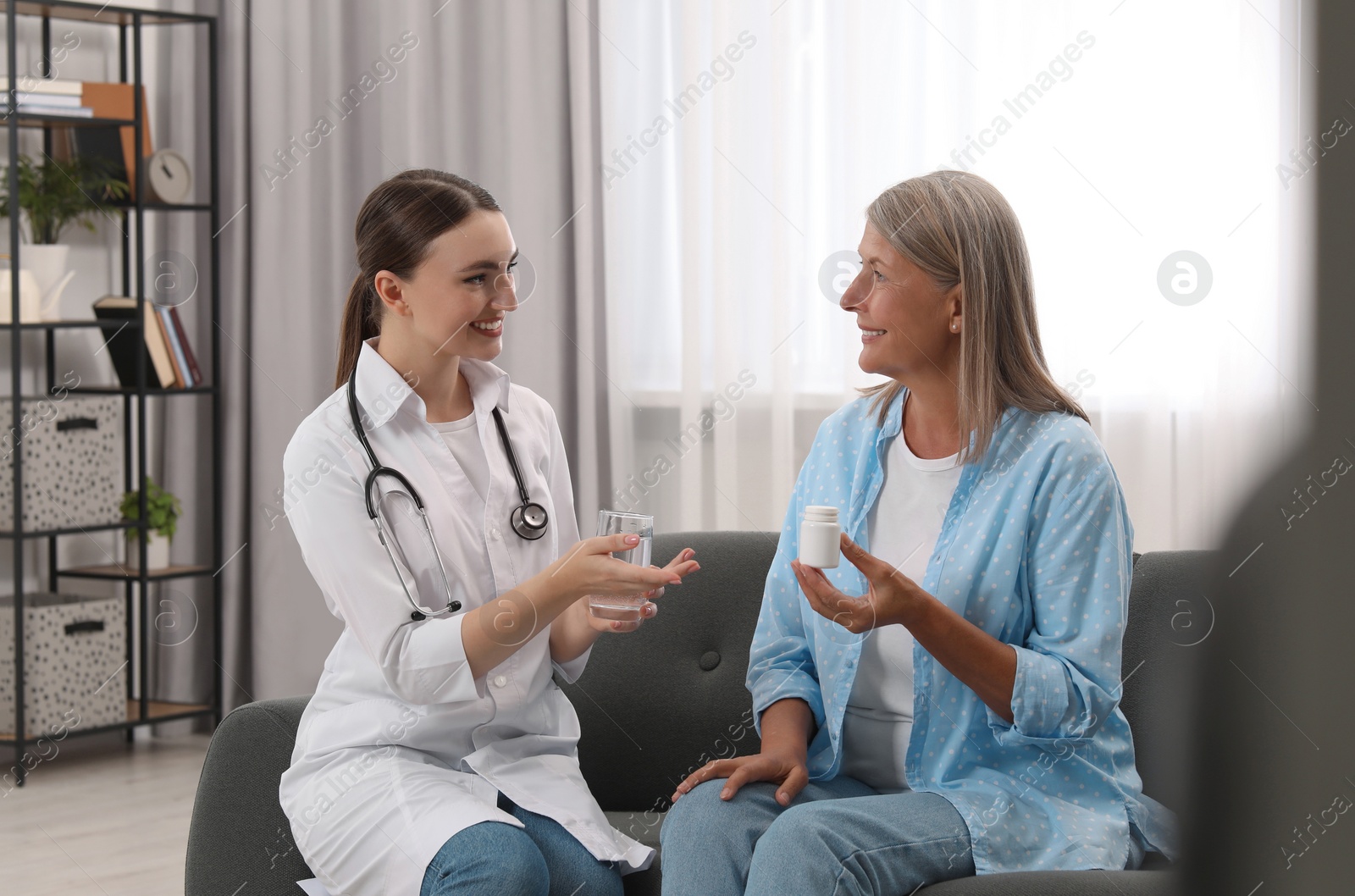 Photo of Young healthcare worker giving glass of water to senior woman with pills indoors