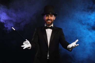Photo of Happy magician holding wand in smoke on dark background