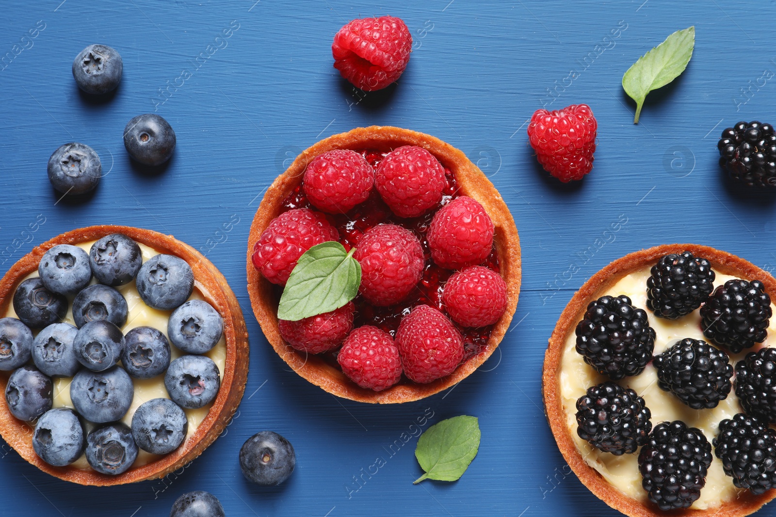 Photo of Tartlets with different fresh berries on blue wooden table, flat lay. Delicious dessert