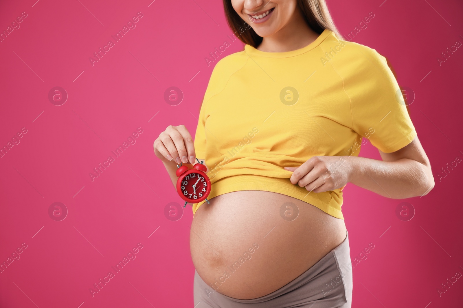 Photo of Young pregnant woman holding alarm clock near her belly on pink background, closeup. Time to give birth