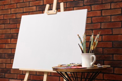 Photo of Wooden easel with blank canvas and different art supplies near brick wall, closeup