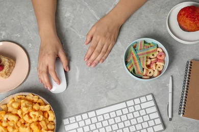 Photo of Bad eating habits. Woman working on computer at grey marble table with different snacks, top view