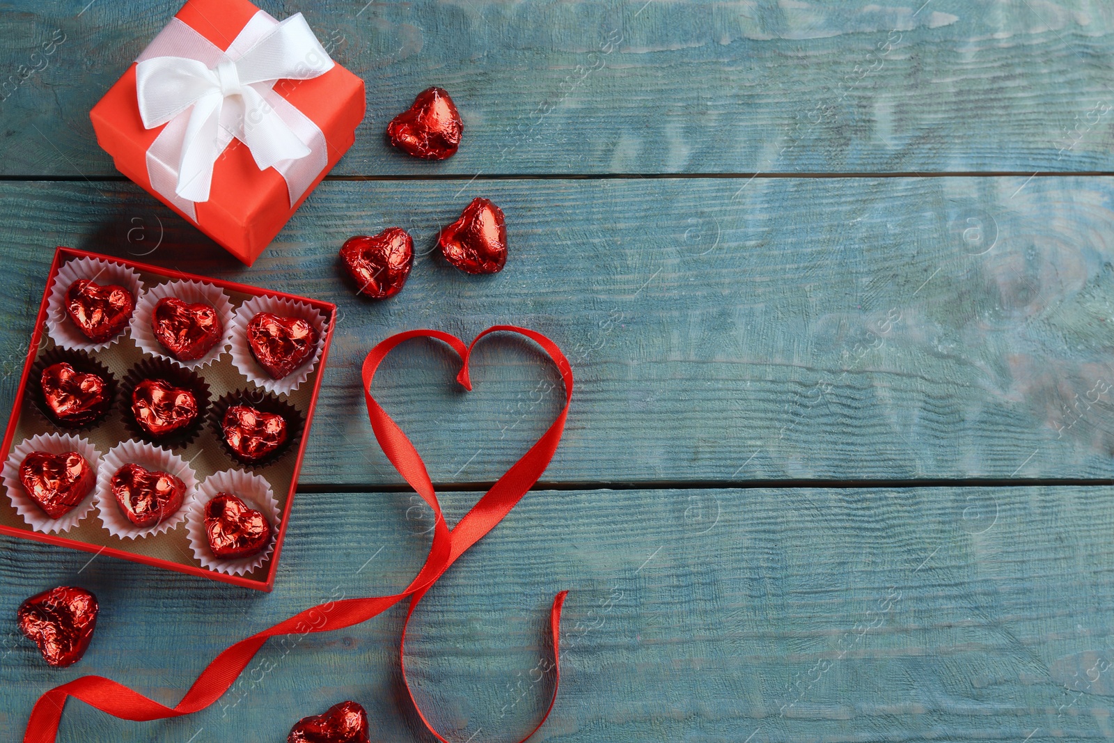 Photo of Heart shaped chocolate candies, gift box and red ribbon on blue wooden table, flat lay with space for text. Valentine's day celebration