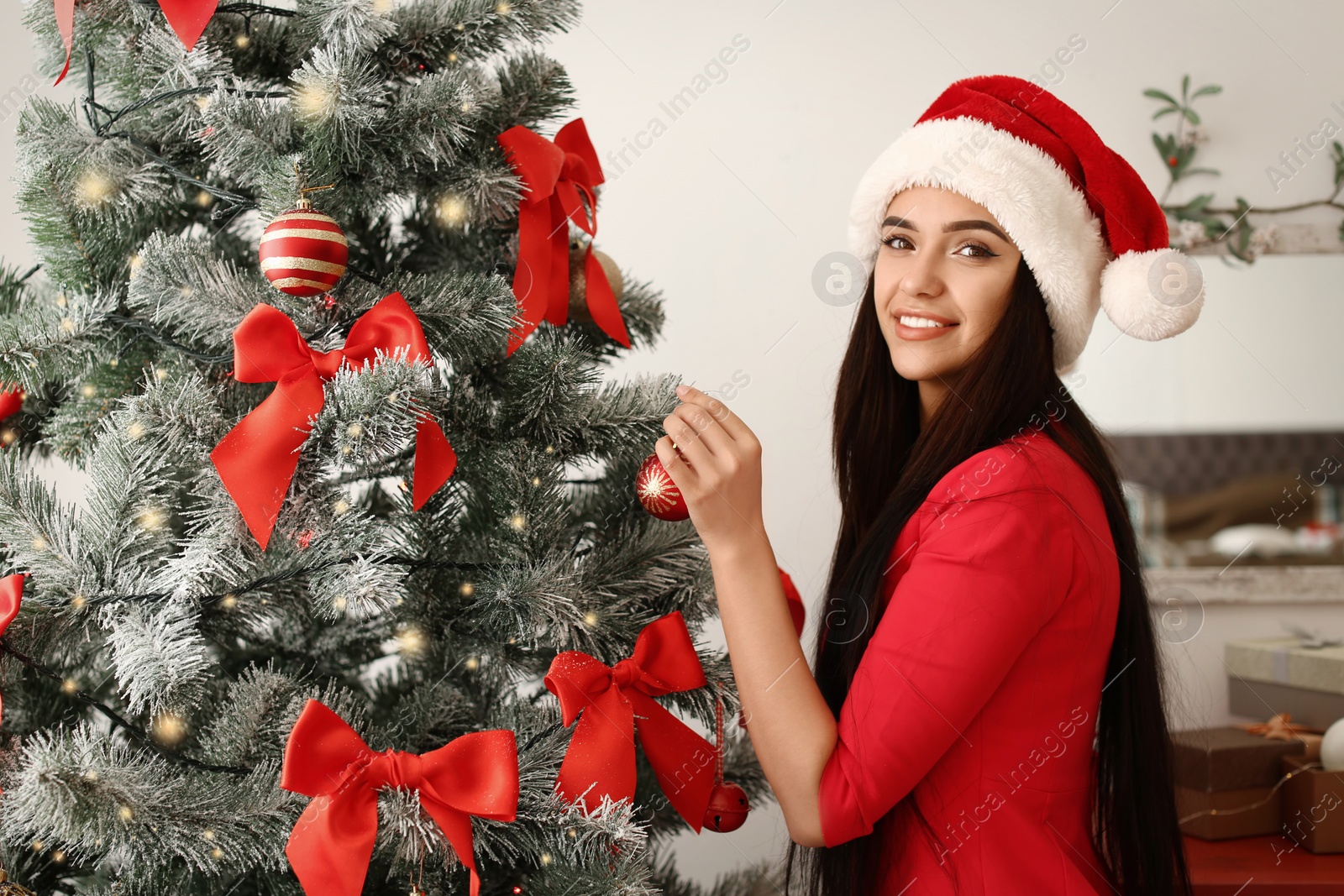 Photo of Beautiful young woman in Santa hat decorating Christmas tree at home