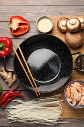 Black wok and chopsticks surrounded by products on color wooden table, flat lay