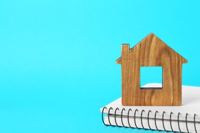 Photo of Wooden house model and notebook on light blue background, closeup with space for text. Mortgage concept